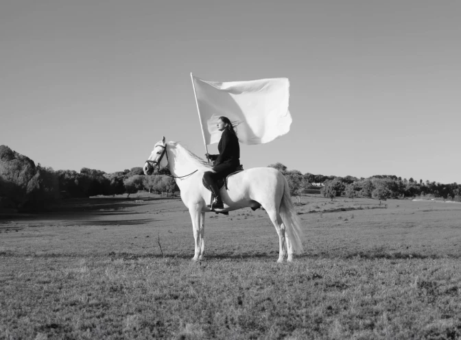 Black and white photo of a woman in black riding a white horse and holding a white flag.