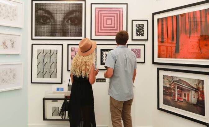 A woman with blonde hair and a man look at art hanging on a gallery wall.