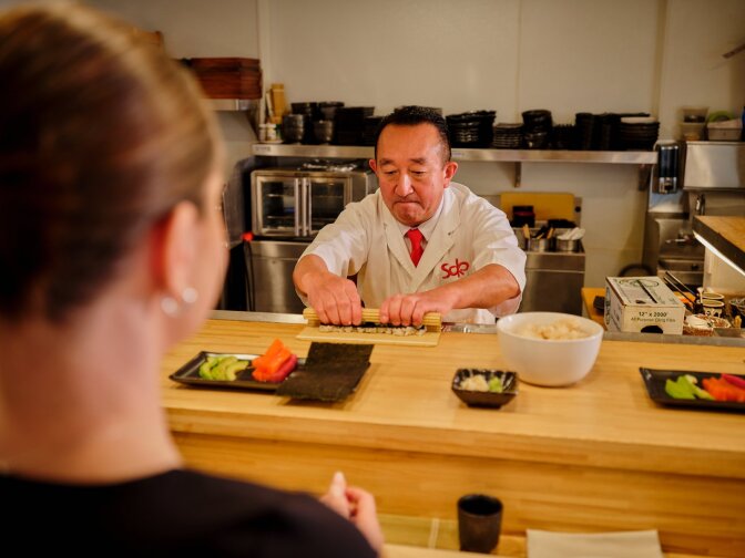 A Japanese man wearing a white chef's coat rolls sushi on a sushi bar, and an out-of-focus back of a person's head looks on in the front left portion of the frame.