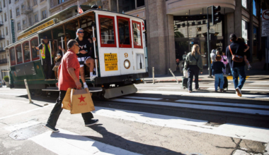 A man carrying Macy's shopping bags crosses a street in front of a historic San Francisco cable car, with other pedestrians walking nearby.