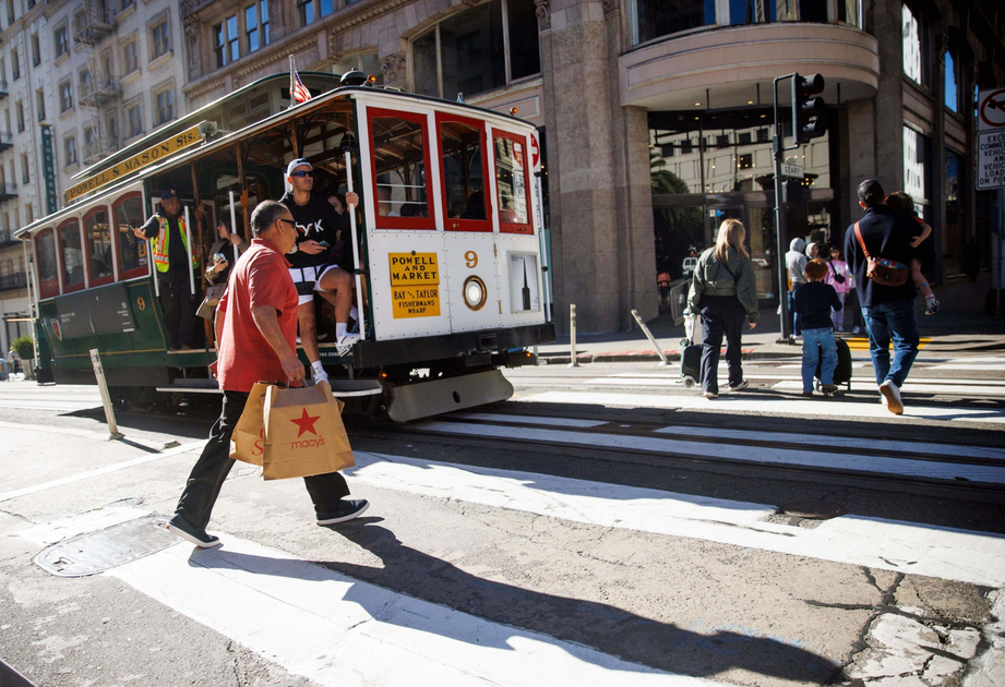 A man carrying Macy's shopping bags crosses a street in front of a historic San Francisco cable car, with other pedestrians walking nearby.