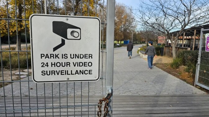 A ign with an illustration of a surveillance camera with the words "Park is under 24 hour surveillance" hangs on a fence. In the distance two people are pictured walking along a concrete pathway.