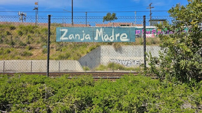 A blue hand painted sign that reads "Zanja Madre (Mother Ditch)" hangs on a chain-link fence. Beyond the fence is a railroad and a concrete wall with graffiti.