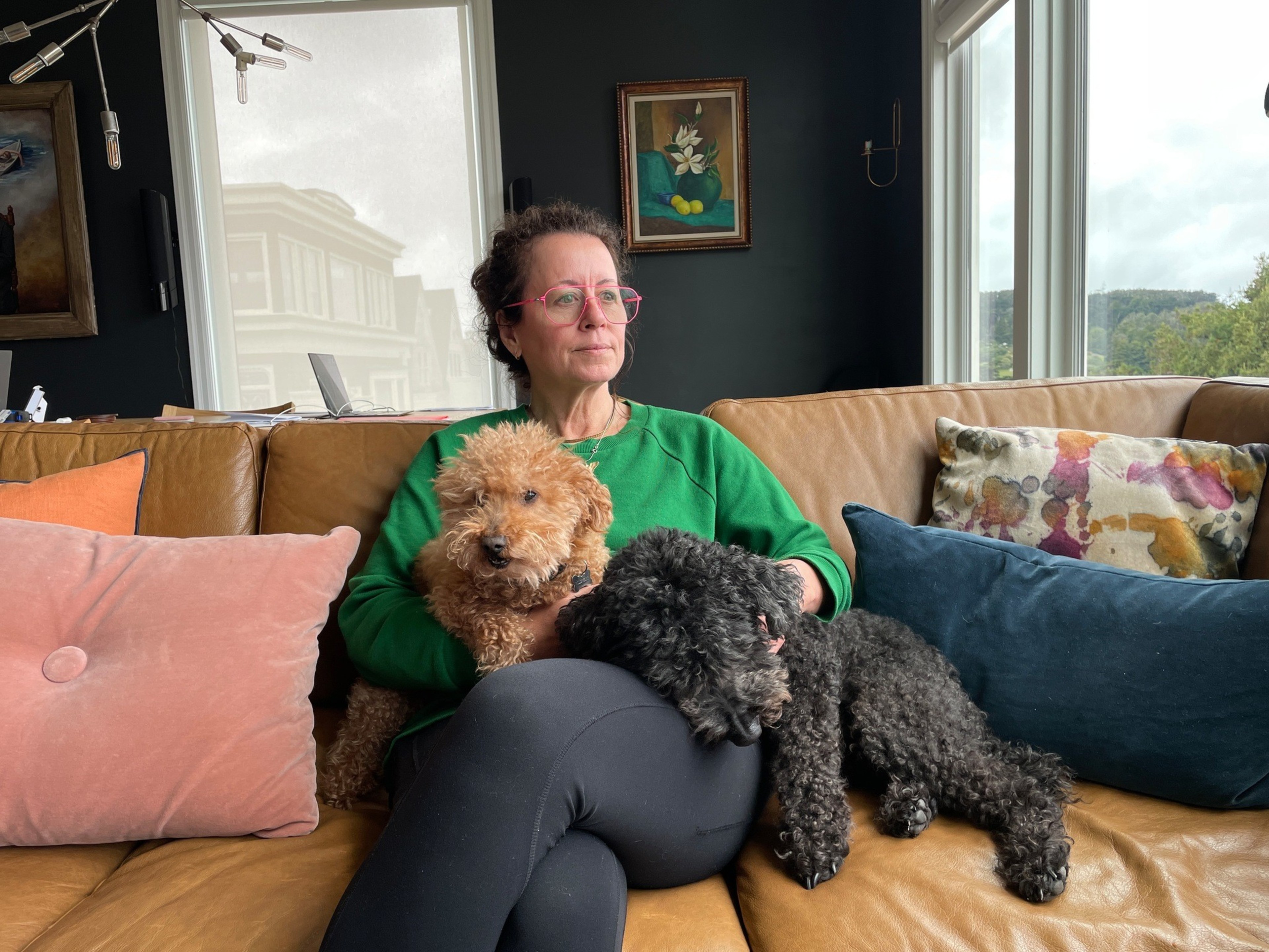 A woman in a green sweatshirt and pink glasses sits on a brown leather couch with two small curly-haired dogs, one tan and one black, resting on her lap.