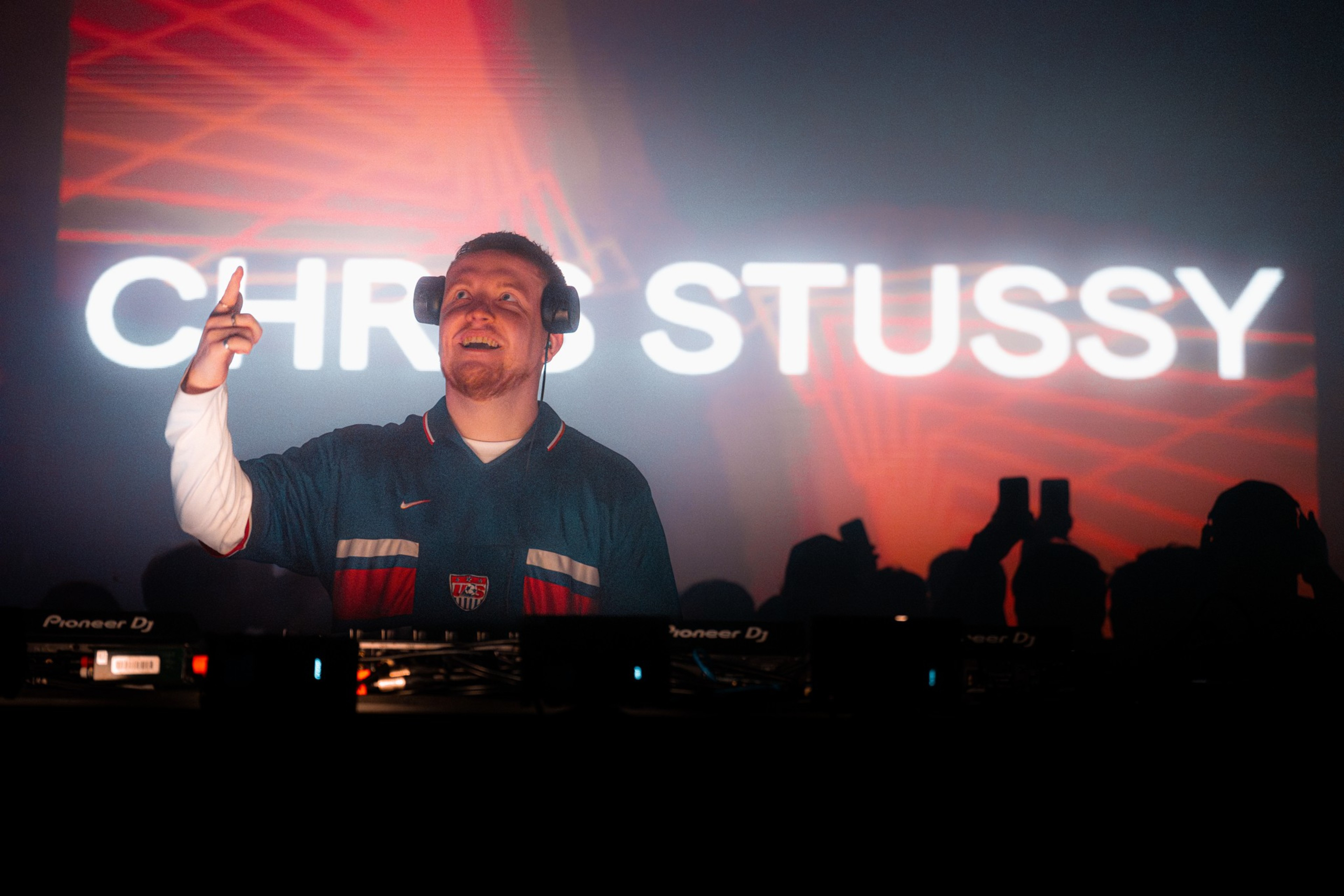 A DJ wearing headphones and a dark jersey points upward while smiling behind a Pioneer DJ booth, with “CHRIS STUSSY” displayed brightly in the background.