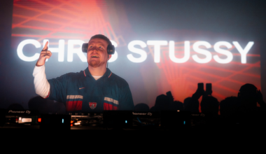 A large crowd gathers inside a warehouse-style club with red and blue lighting, facing a bright screen displaying "CLUB DARC."