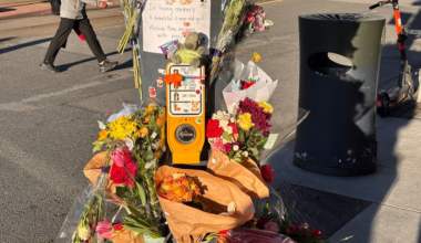A street corner memorial with bouquets of flowers, a teddy bear, and a sign honoring a 2-year-old girl from Mission Bay.