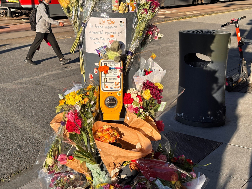 A street corner memorial with bouquets of flowers, a teddy bear, and a sign honoring a 2-year-old girl from Mission Bay.
