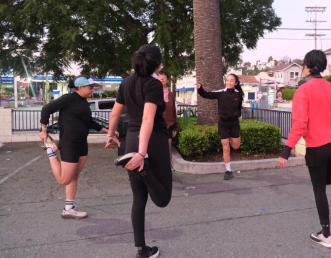A group of people stretch near the corner of a parking lot where a gas station is seen across the street.