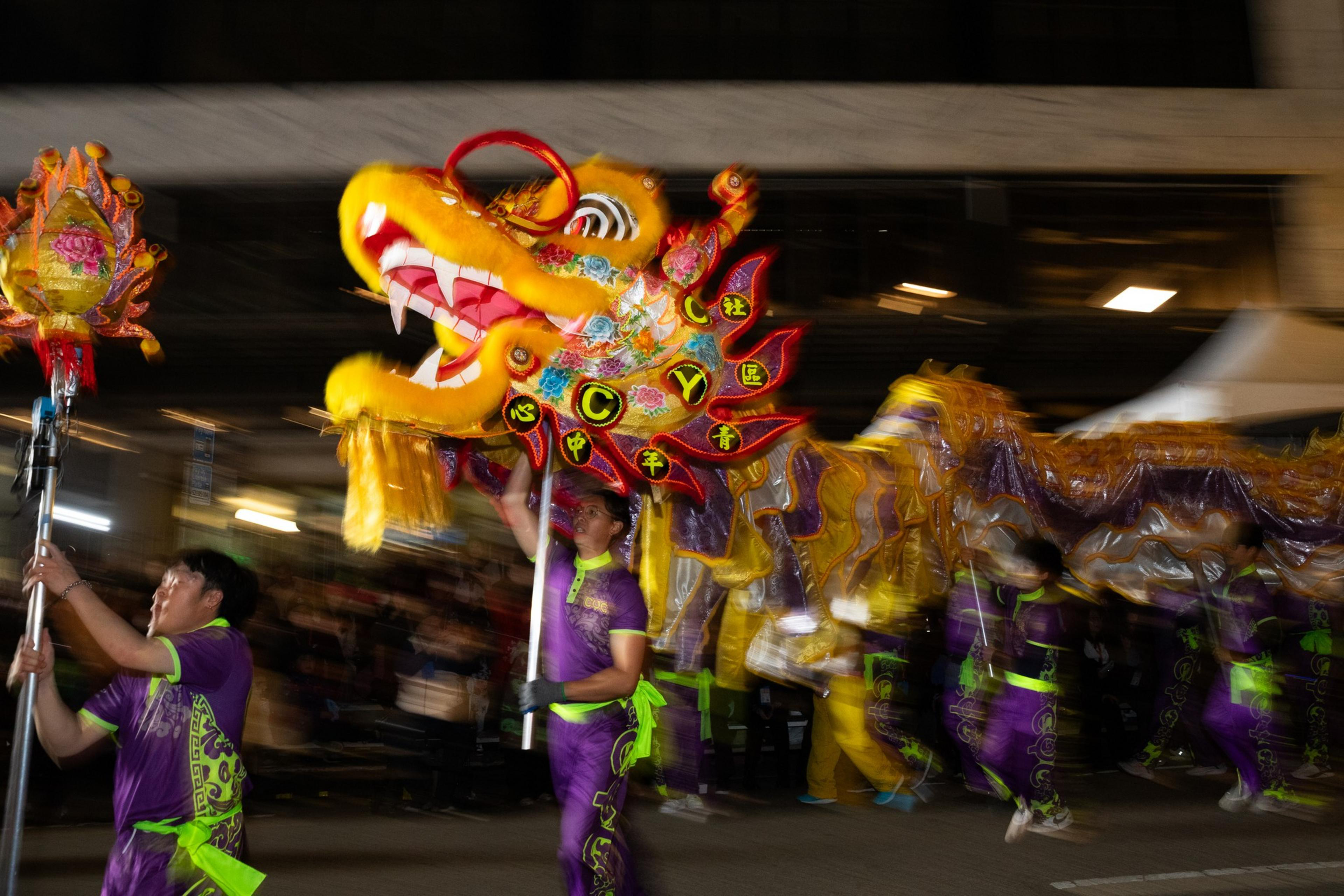 Performers dressed in purple and green carry a vibrant yellow and red dragon costume during a nighttime parade.