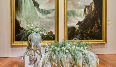 Two large paintings of waterfalls in gold frames are displayed on a wall. In front are two elaborate, contemporary floral arrangements on white pedestals.