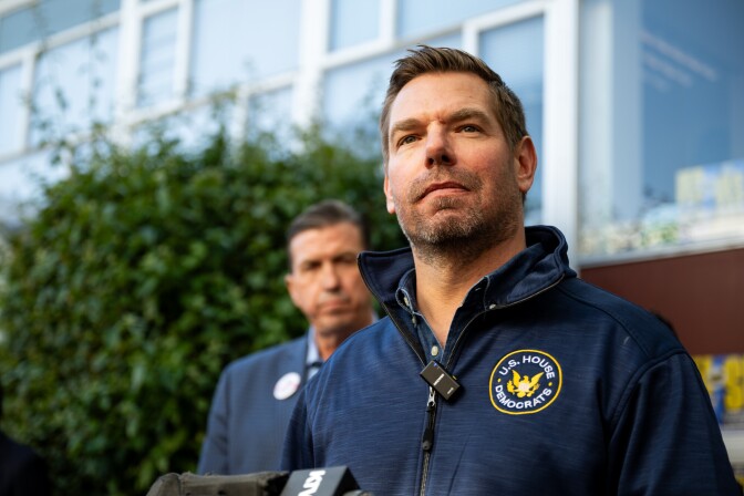 Phot of a man standing outside in front of a blurred building. He is wearing a zippered long sleeve top with a round patch on the left side of his chest that reads "U.S. House Democrats." Another man, wearing a blue suit jacket stands behind him