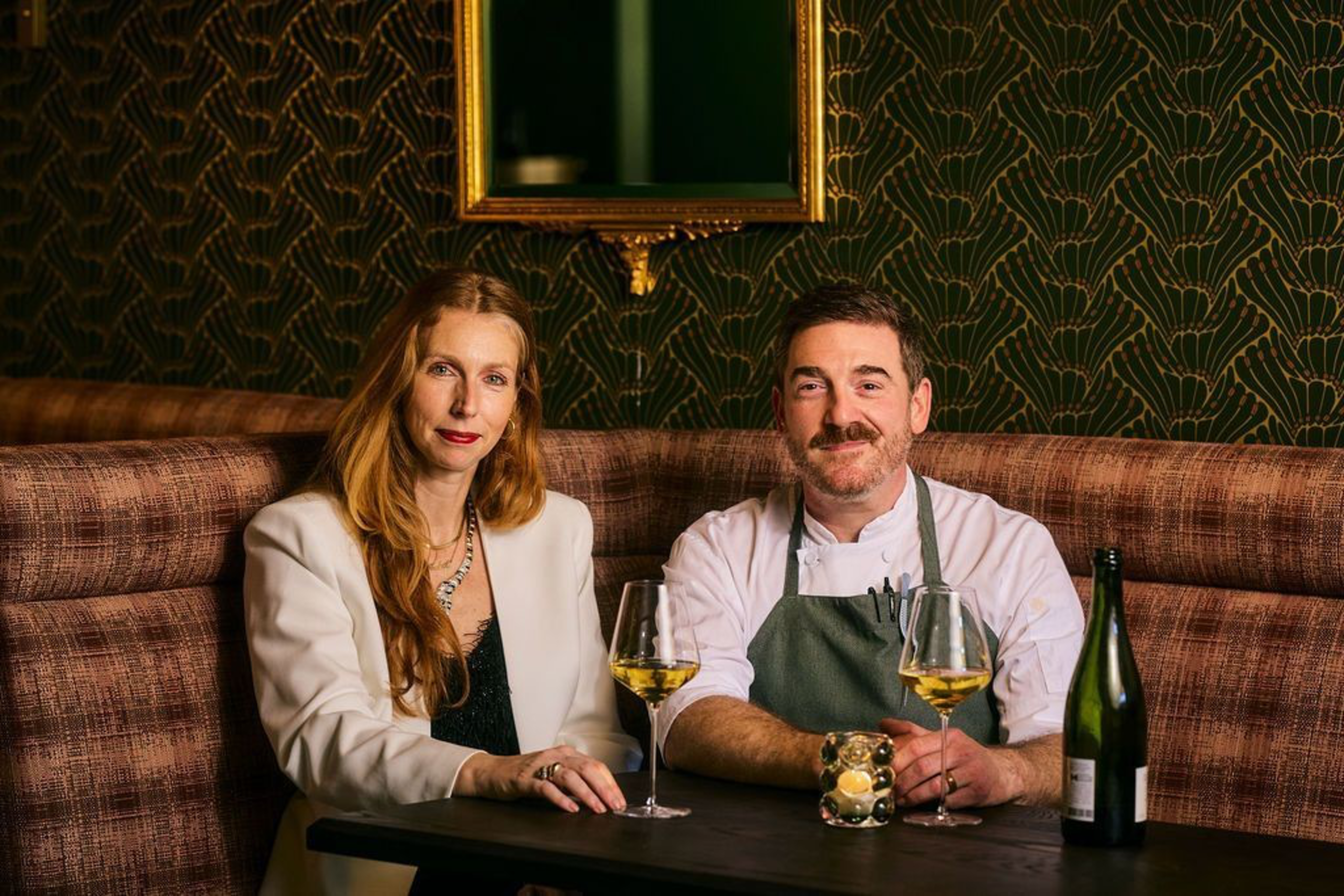 A woman in a white blazer and a man in a chef’s outfit sit at a table with two glasses of white wine and a bottle in a dimly lit, patterned room.