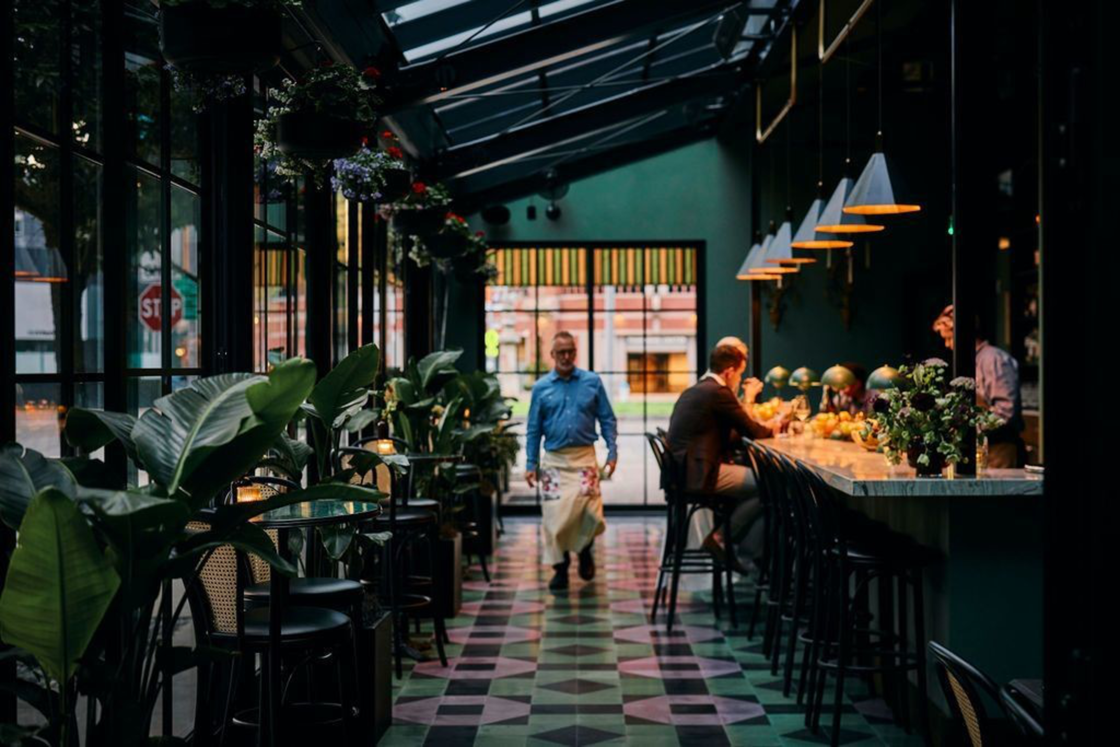 A waiter walks through a dimly lit restaurant with large plants along glass walls, while a man sits at a marble bar counter under hanging lights.