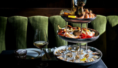 A three-tiered seafood platter features oysters on ice at the bottom, crab legs and lobster in the middle, and shrimp with lemon wedges and sauce on top.