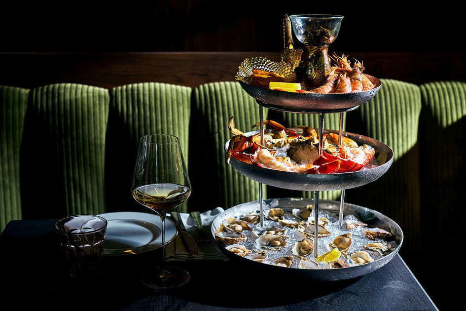 A three-tiered seafood platter features oysters on ice at the bottom, crab legs and lobster in the middle, and shrimp with lemon wedges and sauce on top.