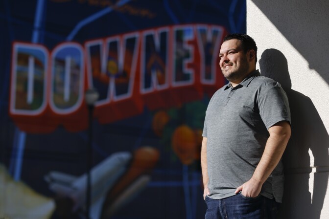 A man with light skin tone and short, dark hair stands against a wall with his hands in his pockets. Behind him, an out-of-focus mural reads "DOWNEY."