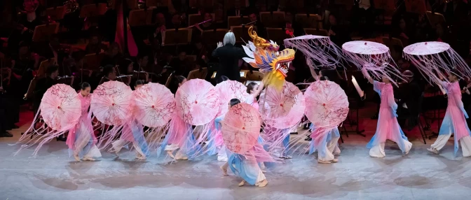 A line of dancers dressed in pink, holding pink umbrellas, dancing across a stage while a symphony plays in the background. 