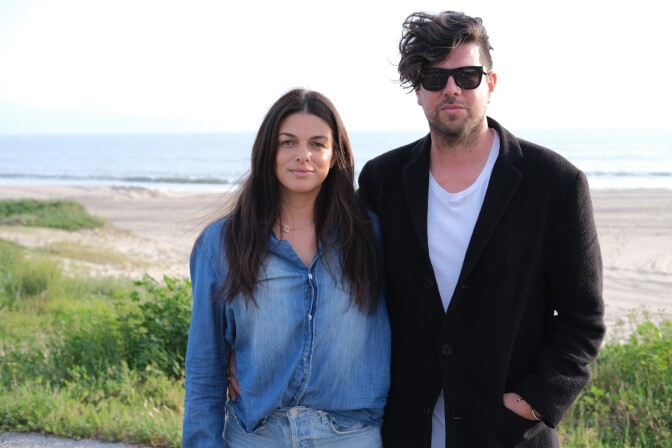  Giorgi Antinori, a woman with light skin tone, and her husband, a man with light skin tone, stand at the edge of Will Rogers State Beach. 