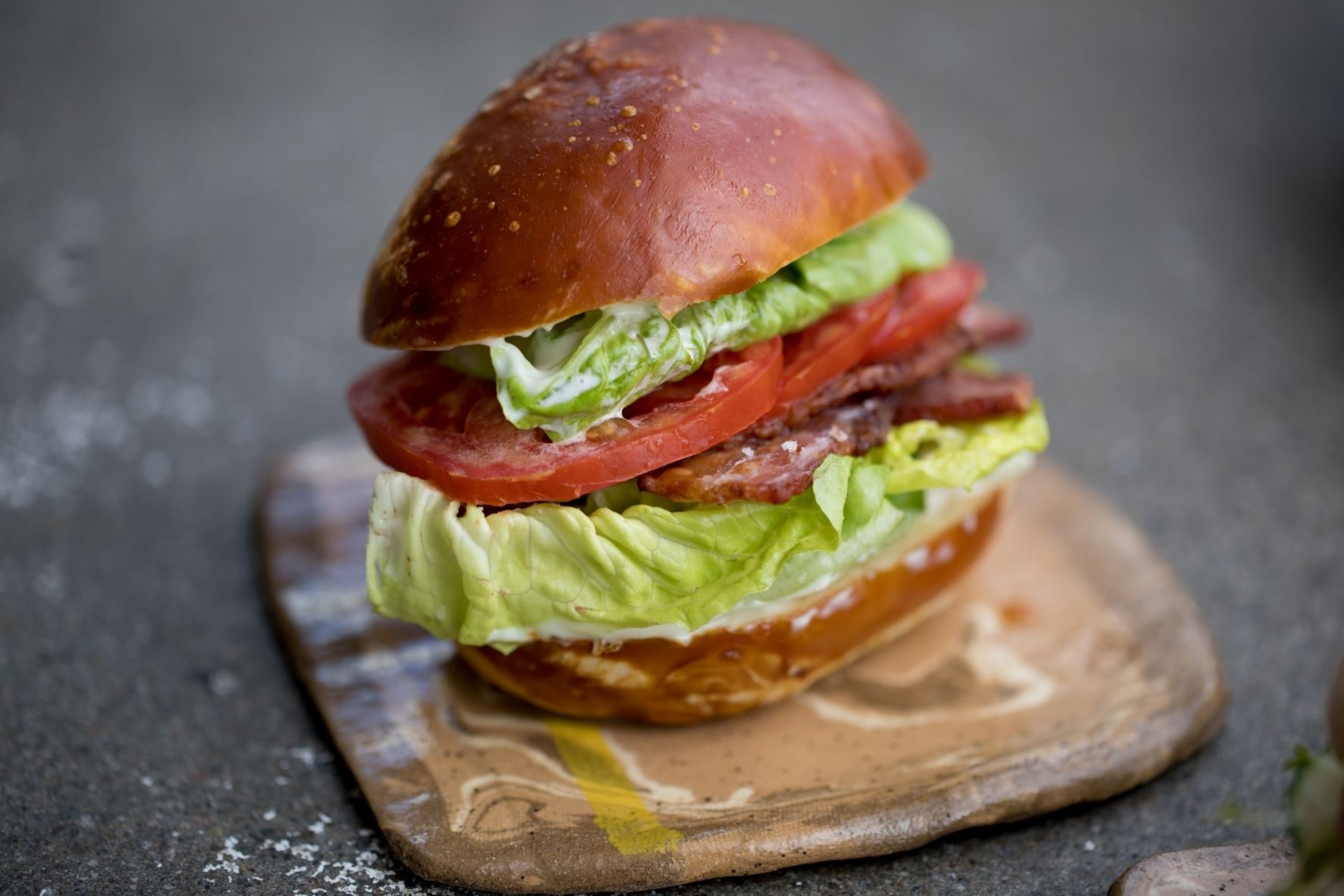 A sandwich with crispy bacon, fresh lettuce, and sliced tomato is placed on a square rustic wooden board.