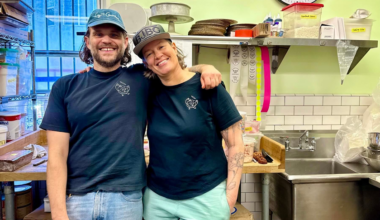Two smiling people in black shirts and caps stand close with arms around each other in a small kitchen with shelves, containers, a sink, and a clock showing 12:15.