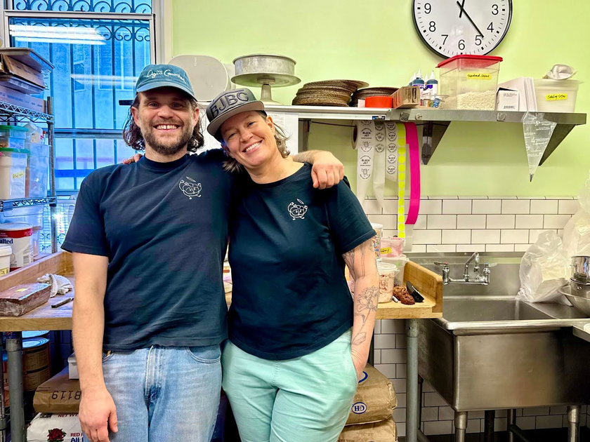 Two smiling people in black shirts and caps stand close with arms around each other in a small kitchen with shelves, containers, a sink, and a clock showing 12:15.