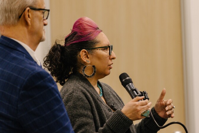 A woman with blotch of pink-colored hair speaks into a microphone as an older white man looks on.