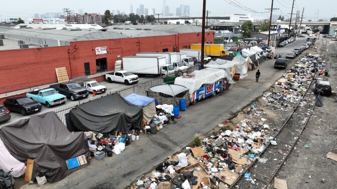 An aerial view of a street with the downtown L.A. skyline in the distance. A set of red buildings are to the left, in front of a line of tents, canopies and shelters in a homeless encampment. Large piles of trash can be seen on the other side of the encampment along train tracks.