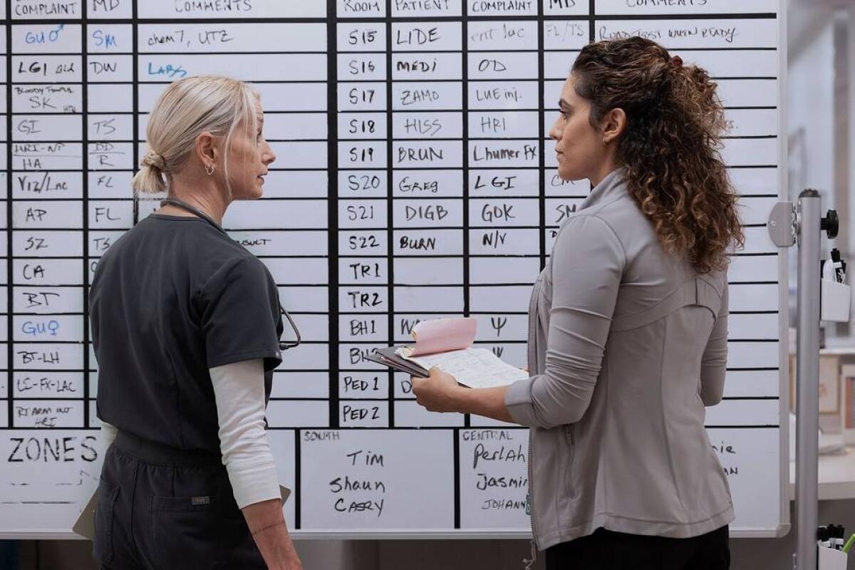 A woman in blue scrubs stands in front of a white board looking at a woman in a mauve jacket holding a clipboard.