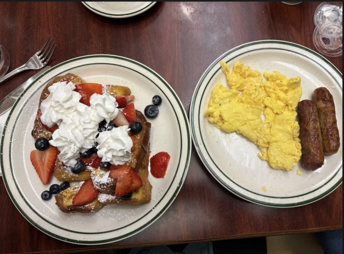 Two white plates side by side; the one on the left has french toast covered with strawberries, blueberries and whipped cream; the one on the right has scrambled eggs and sausages