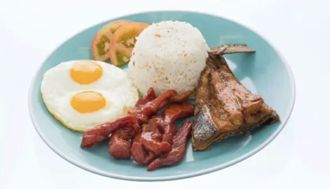 a pale blue plate contains two eggs, rice, tomatoes, pieces of cured meat and a slab of fish