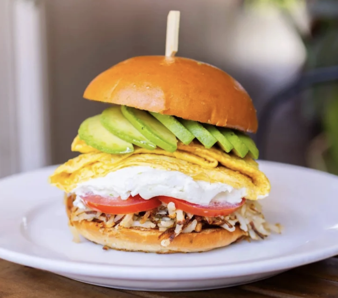 A burger roll which contains a folded omelette, cream cheese, hash browns, tomatoes and avocado slices