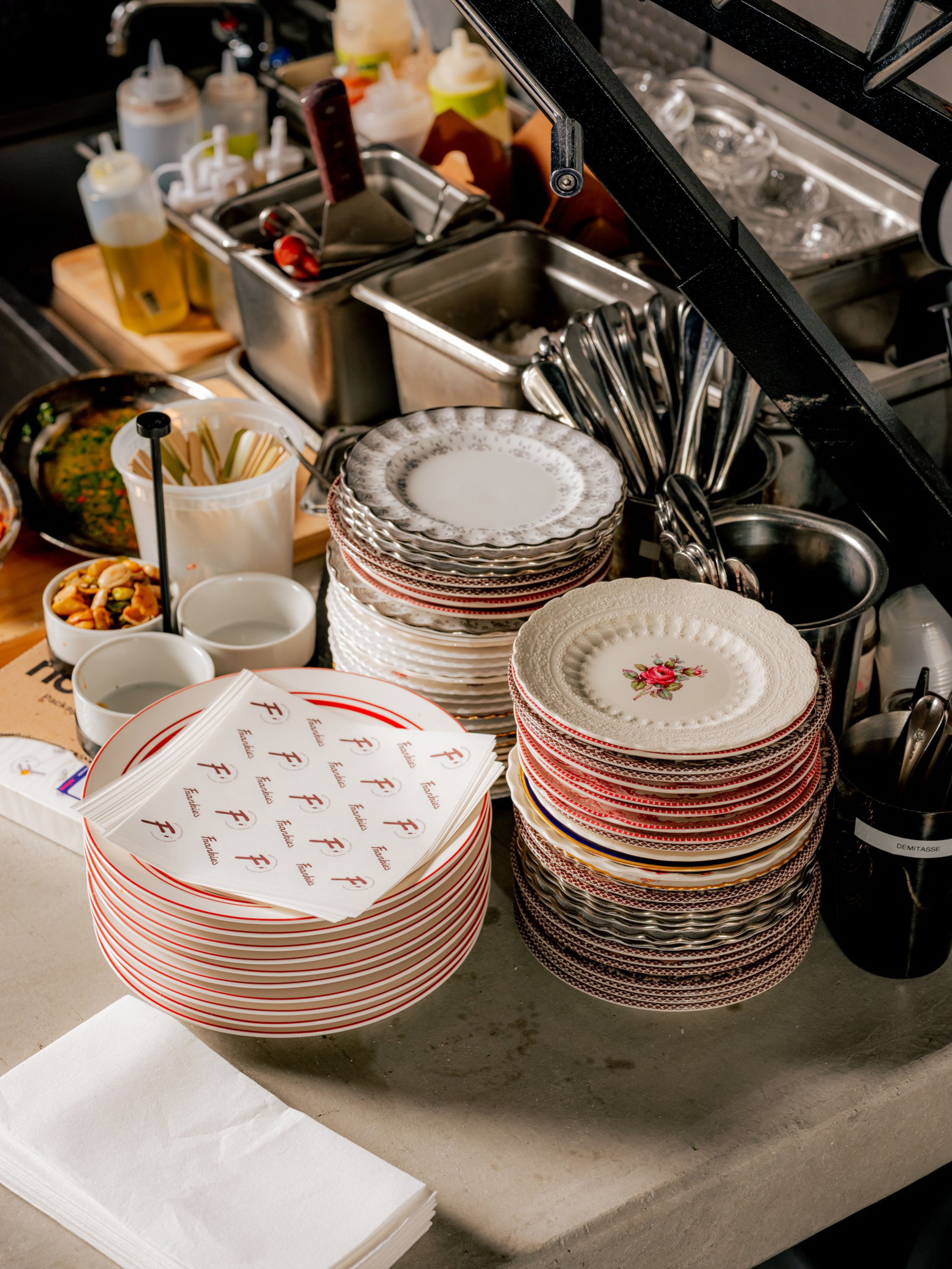 Stacks of various patterned plates, utensils, napkins, and condiment bottles are arranged on a kitchen counter near food prep containers.