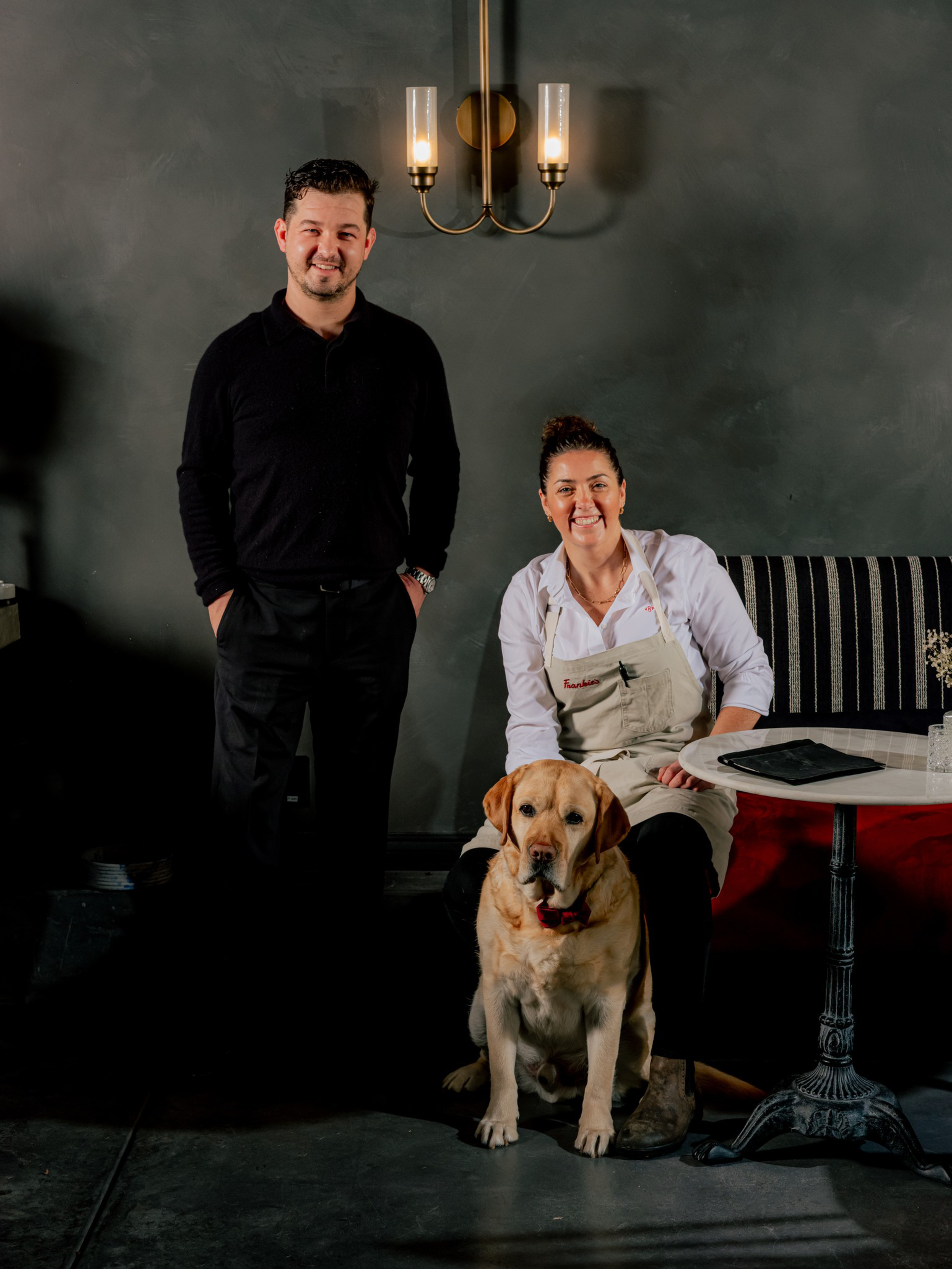 A man in black stands beside a woman in a white shirt and apron sitting on a bench, with a large yellow Labrador sitting in front of them.