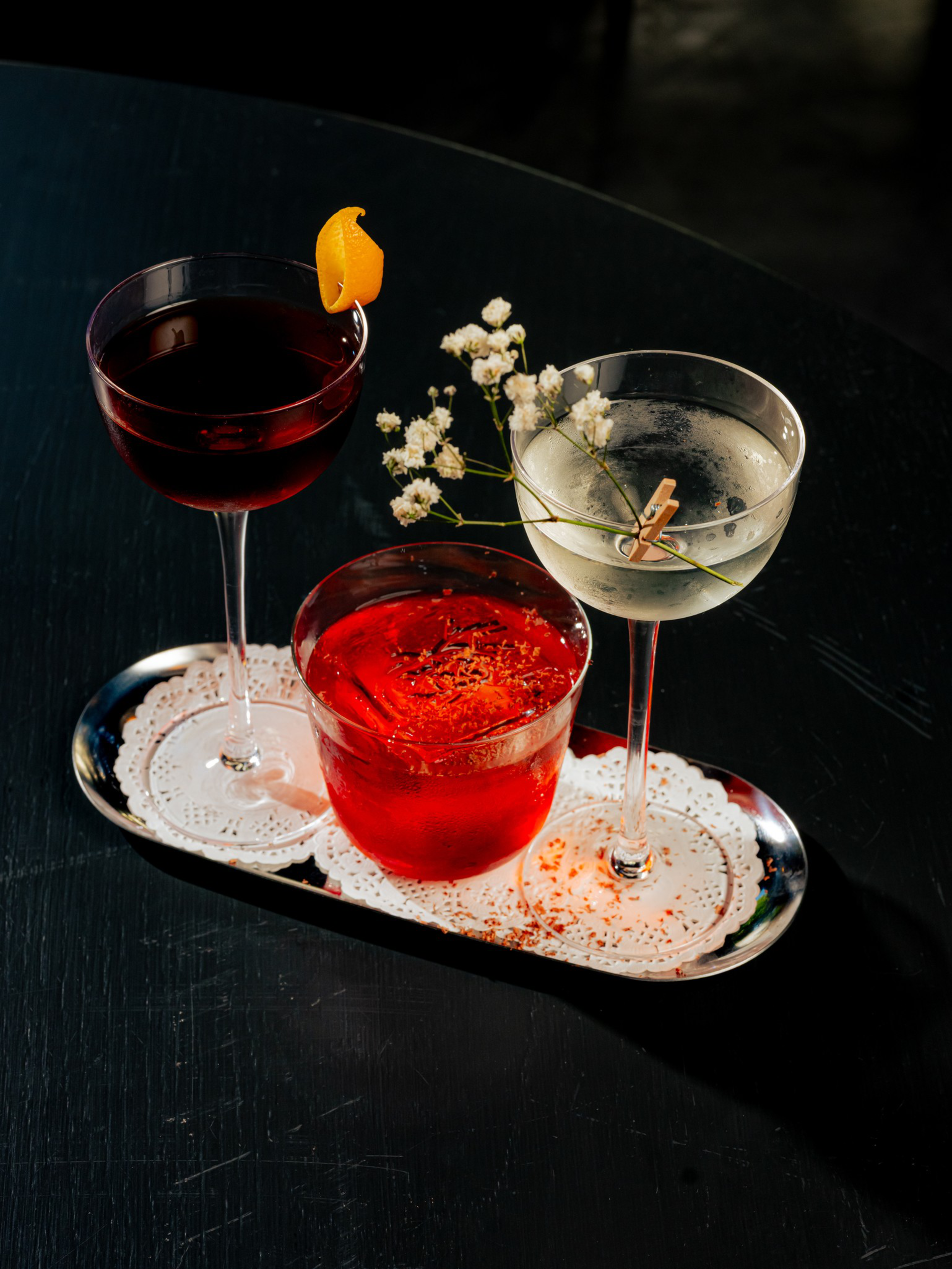 Three cocktails—a dark red drink with an orange peel, a bright red drink in a short glass, and a clear drink with a white flower garnish—sit on a silver tray.