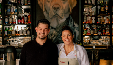 A man and woman smile behind a bar lined with liquor bottles and glasses, with a large portrait of a dog dressed in clothing on the wall behind them.