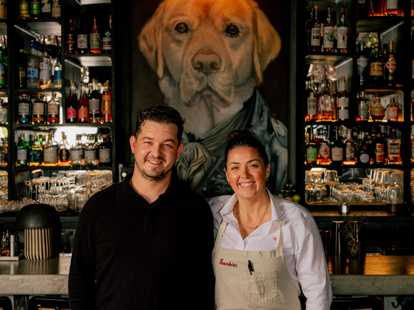 A man and woman smile behind a bar lined with liquor bottles and glasses, with a large portrait of a dog dressed in clothing on the wall behind them.