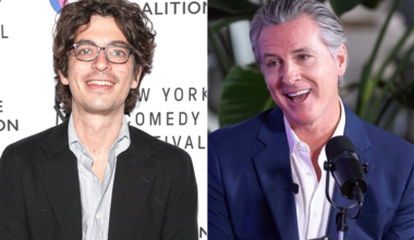 A man with curly dark hair and glasses wears a black jacket and striped shirt, while a smiling man with gray hair in a blue suit speaks into a microphone.