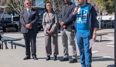 Four people stand outdoors on a concrete platform; one man in a blue outfit speaks into a microphone while the others listen attentively.