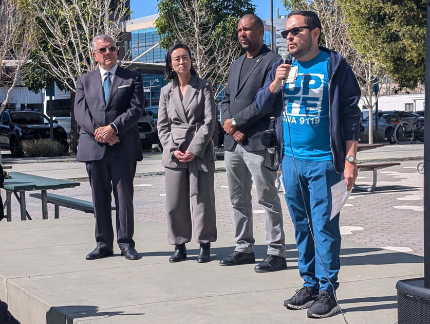 Four people stand outdoors on a concrete platform; one man in a blue outfit speaks into a microphone while the others listen attentively.