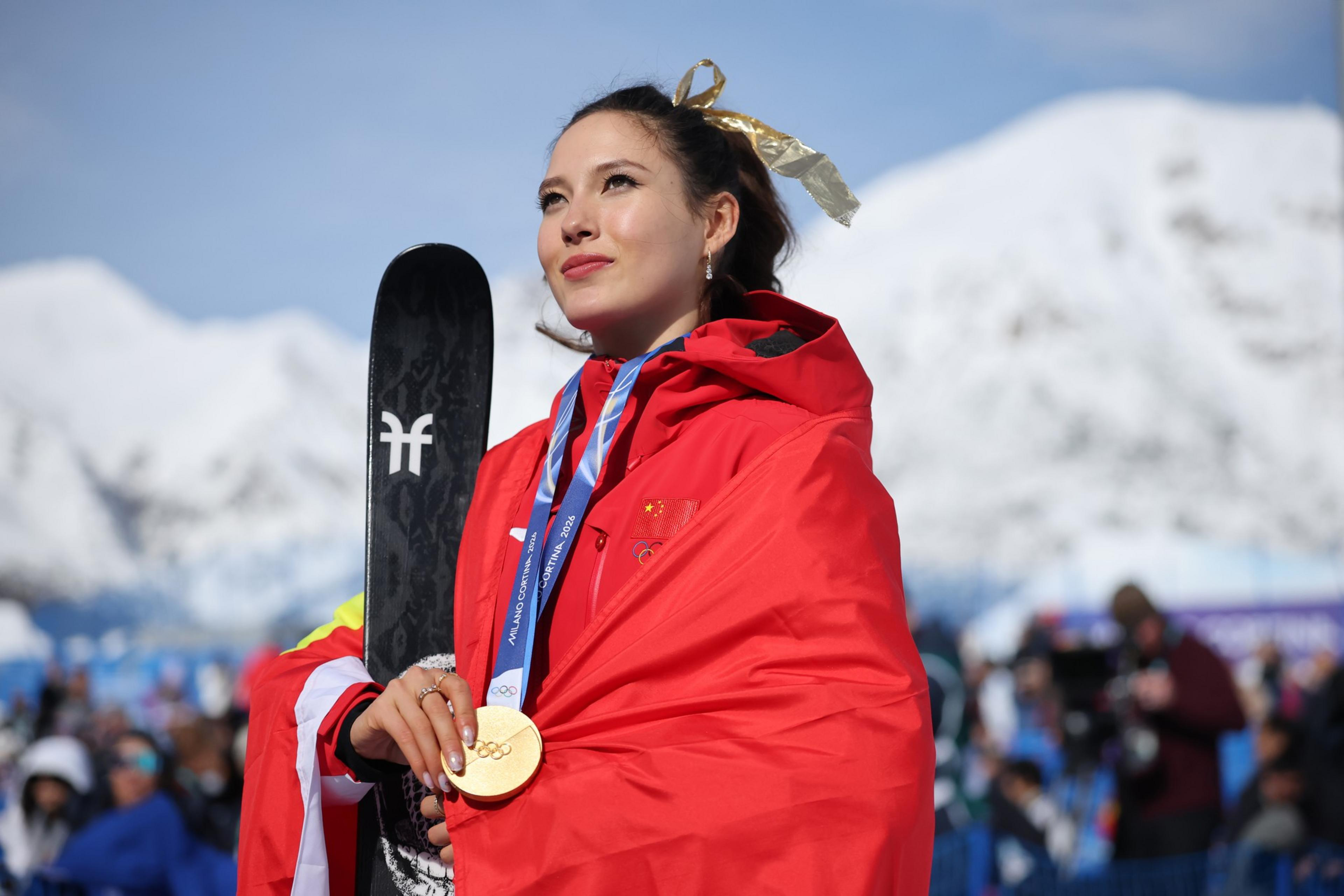 A woman wrapped in a red flag holds a ski and wears a gold Olympic medal with snowy mountains blurred in the background.