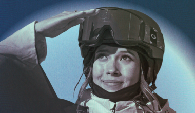 A person wearing a ski helmet and jacket looks up with a slight smile, shielding their eyes with one hand against the sun.