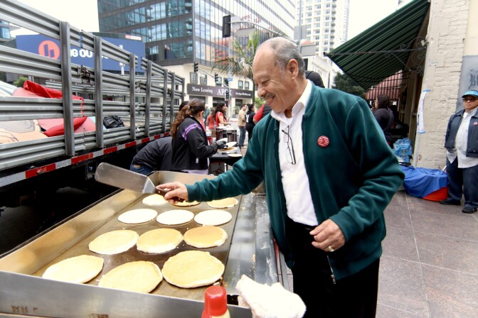 An older Latino man is flipping pancakes on a grill set-up on a sidewalk. The man is smiling and wearing a dark green jacket over his white shirt and black pants. Several other people can be seen on the sidewalk behind him.