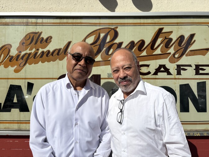 Two older Latino men in white long-sleeve button down shirts are standing shoulder to shoulder in front of a large sign that reads "The Original Pantry Cafe." The man on the left is wearing sunglasses, while the man on the right has glasses hanging from his shirt collar.