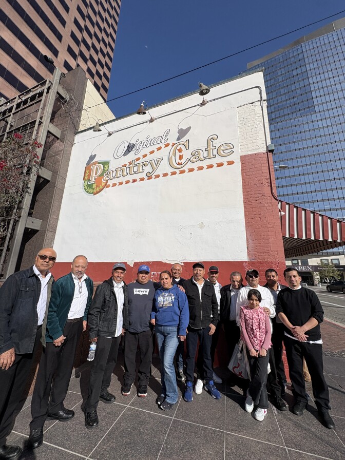 A group of men and women are standing in a group and posing in front of the side of a large building. The white and red wall facing the camera reads "Original Pantry Cafe"