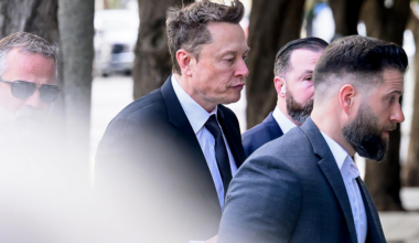 Four men in suits walk closely together outdoors, with one man in the center having distinctive messy hair and a serious expression.