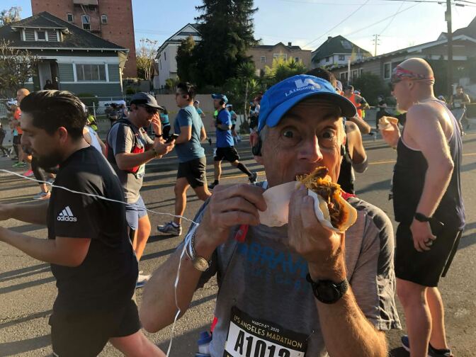 A man in marathon gear eating a chili cheese dog. Runners are everywhere on the street behind him.