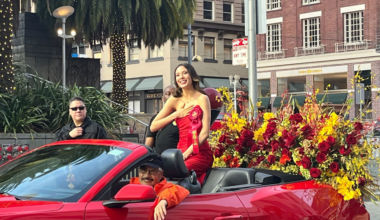 A woman wearing a red dress and a ribbon sash smiles while sitting in a red convertible filled with red and yellow flowers during a parade.
