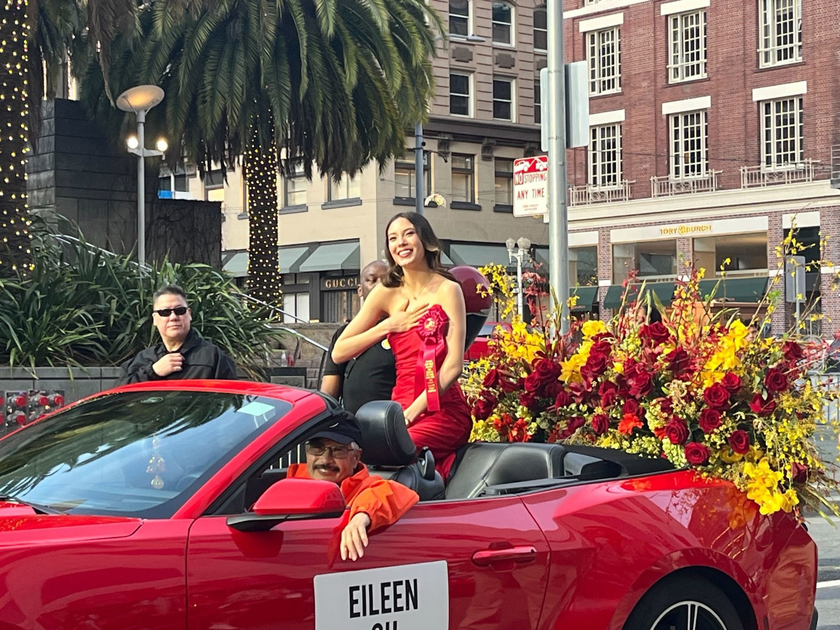 A woman wearing a red dress and a ribbon sash smiles while sitting in a red convertible filled with red and yellow flowers during a parade.