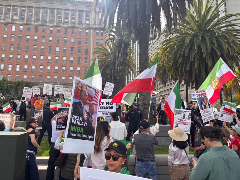 People hold Iranian flags and protest signs advocating against Iran's regime, with palm trees and a large building in the background.